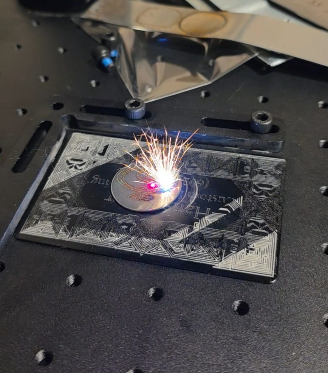 Fiber laser mid-engrave on a coin, throwing sparks, on a custom-engraved fixture plate
